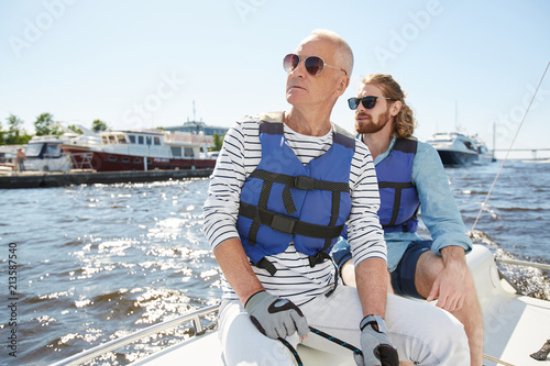 Serious relaxed tourists in fashionable sunglasses and life jackets looking around while sitting on sailboat deck during sailing tour