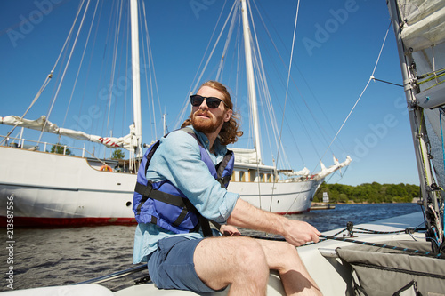 Serious carefree young bearded man in sunglasses operating sail with rope and looking around during sailing travel