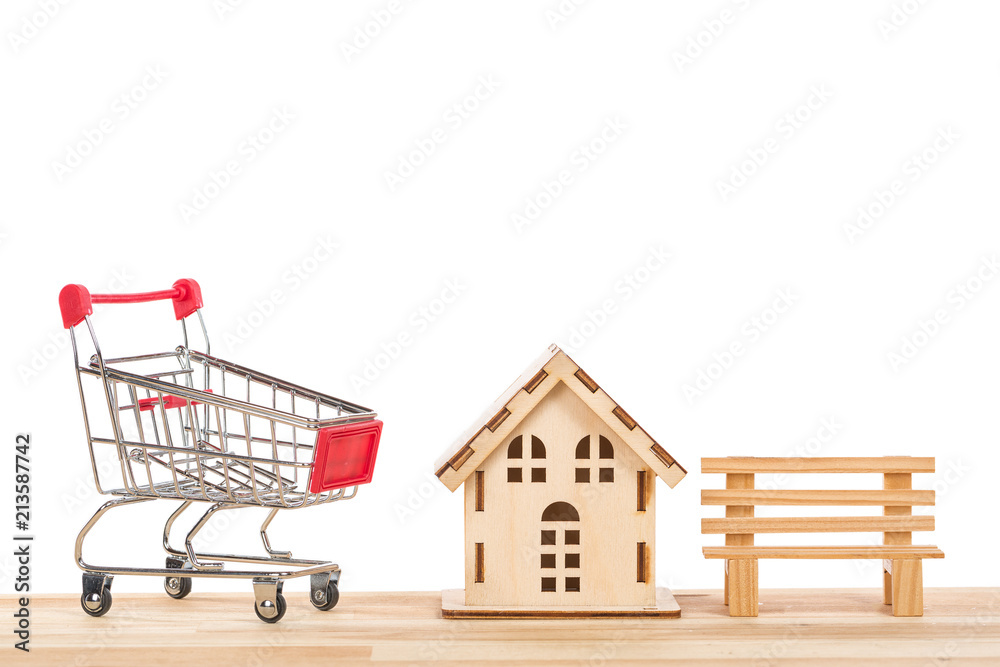 Shopping cart trolley with little wooden house and bench on a table against white isolated background. Buying a house or apartment concept.