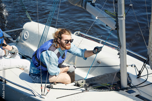 Serious focused young bearded sportsman in sunglasses using ropes while directing sailboat during competition