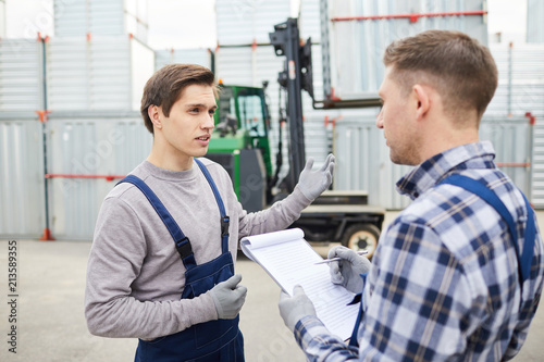 Misunderstanding young cargo employee in workwear gesturing while asking foreman about loading and explaining characteristics of forklift
