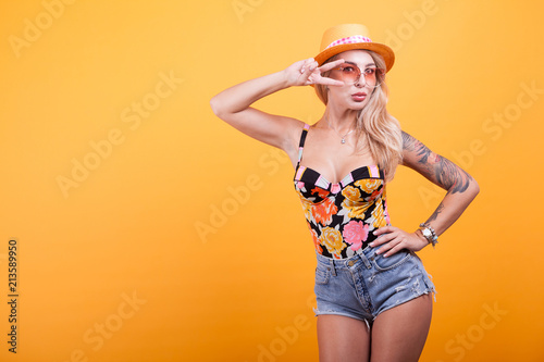 Beautiful young woman making hand signs with sunglasses and hat over yellow background in studio photo