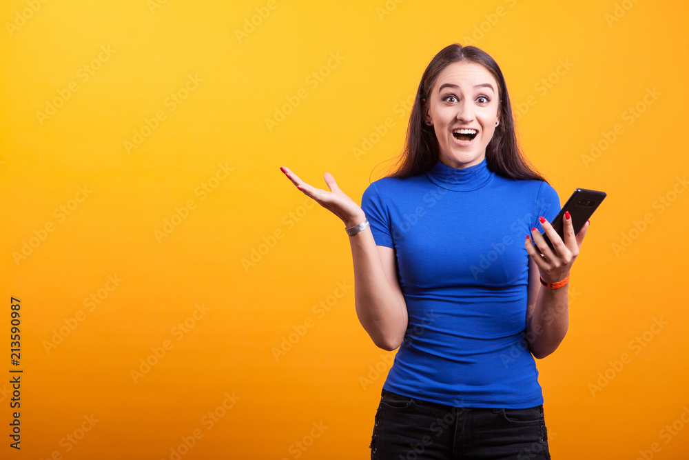 Surprised and amazed girl holds her phone in studio on yellow background Surprised and amazed girl holds her phone in studio on yellow background