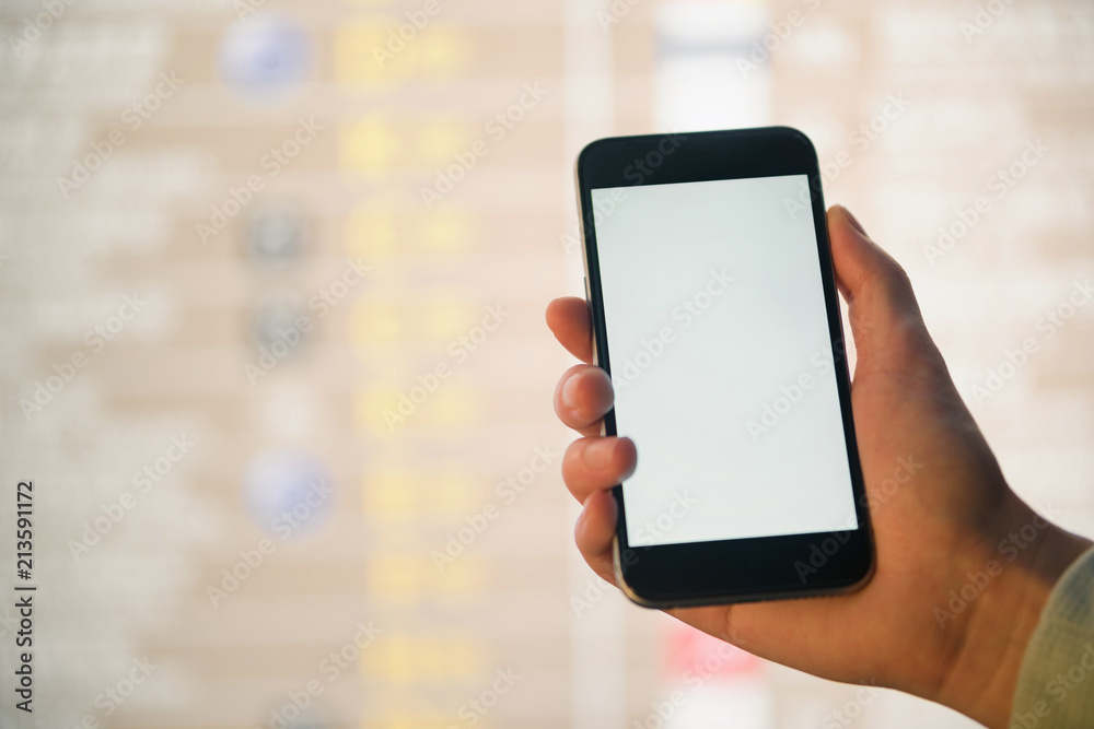 Mockup image of female hands holding black mobile phone with blank white screen over flight board in airport terminal