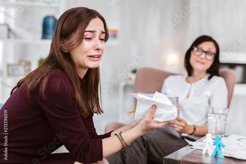 Tears. Sad unhappy young person sitting at the psychological sessional and crying while talking about her problems photo