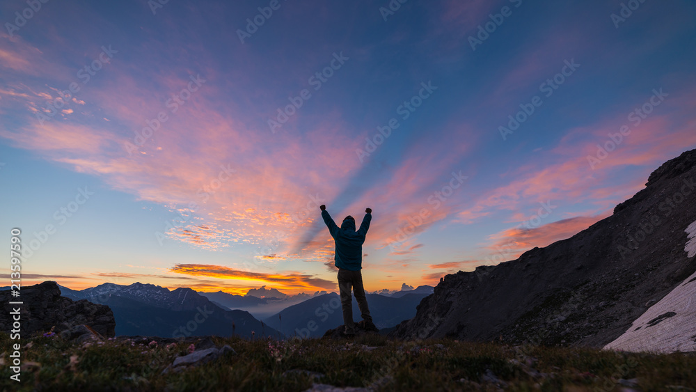 Man standing on mountain top raising arms, sunrise light colorful sky scenis landscape, conquering success leader concept.