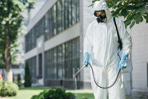 pest control worker in uniform and respirator spraying pesticides on street with sprayer