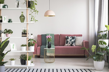 Lamp above table with flowers in front of red sofa in white living room interior with plants. Real photo