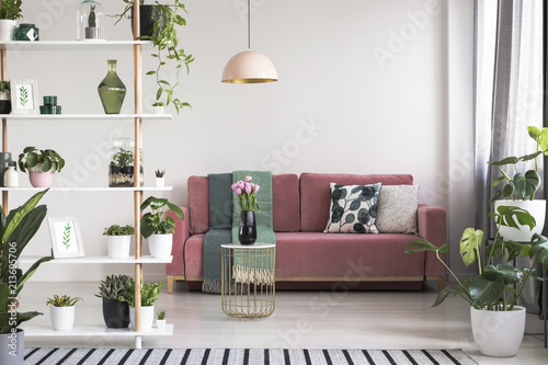 Lamp above table with flowers in front of red sofa in white living room interior with plants. Real photo