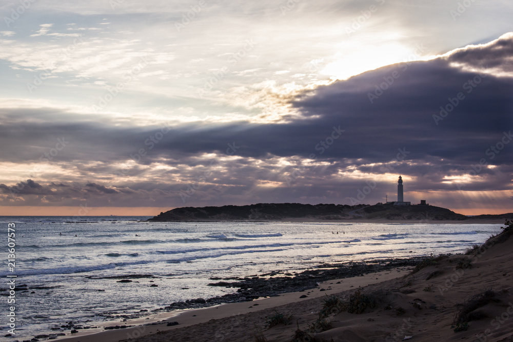 Obraz premium Great panorama of group of surfers enjoying waves on stunning sunset with Trafalgar lighthouse in Cadiz province, Spain. Summer vacation, travel destination concepts