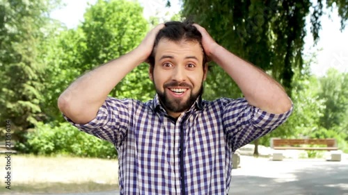 Amazed man is looking on camera. He is smiling cheerful. Guy touches hair and grabbed it with hands. Then he is waving with hands and touching head. Guy lookes down, touches beard and loooks straight. photo