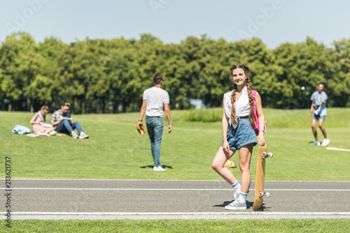 teenage girl with skateboard smiling at camera while friends spending time behind in park