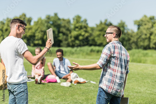 happy teenage boys smiling each other and holding book while friends studying behind in park © LIGHTFIELD STUDIOS