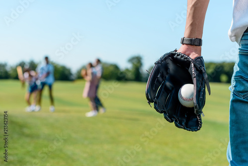 cropped shot of teenager in baseball glove holding ball while playing with friends in park