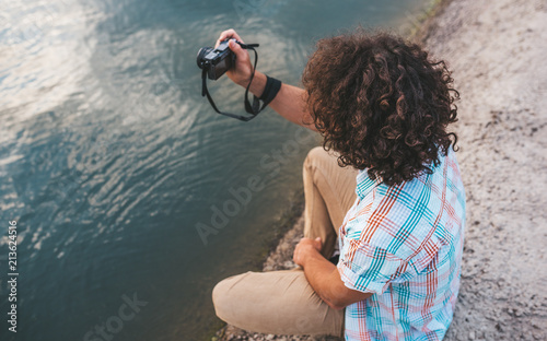 Side view of young male with curly hair checking photos of nature on his digital camera. Handsome man wears casual shirt, with digital camera posing next to the lake. People, travel, lifestyle