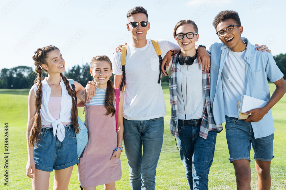 happy multiethnic teenage friends standing together and smiling at camera in park