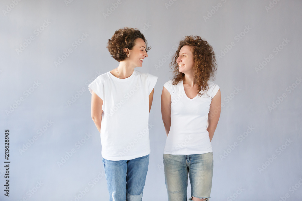 Studio shot of two European sisters with curly hair, wearing white t-shirts and jeans, smiling happily, looking at each other, posing at gray wall. People and friendship concept Studio shot of two European sisters with curly hair, wearing white t-shirts and jeans, smiling happily, looking at each other, posing at gray wall. People and friendship concept