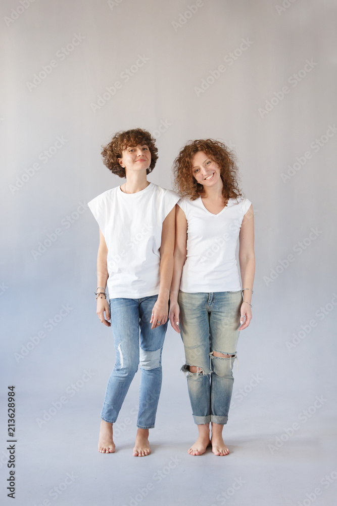 Vertical studio shot of two curly redhead twins sisters or friends stand next to each other against concrete gray wall. Care of elder sister about the younger. Family portrait, friendship concept Vertical studio shot of two curly redhead twins sisters or friends stand next to each other against concrete gray wall. Care of elder sister about the younger. Family portrait, friendship concept
