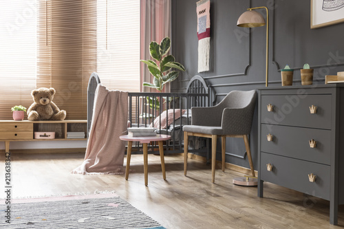 Real photo of a cot standing next to an armchair, lamp and cupboard in dark and classic baby room interior photo