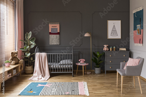 Real photo of a grey crib standing next to a pink stool, a lamp and cupboard in grey baby room interior also with armchair, rug and posters