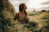Young naked woman sits near haystack.