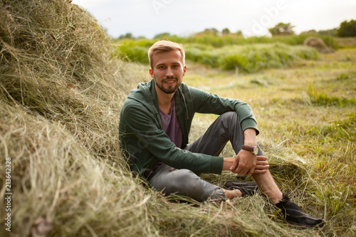 Bilde på lerret Young smiling man sits near haystack on meadow.