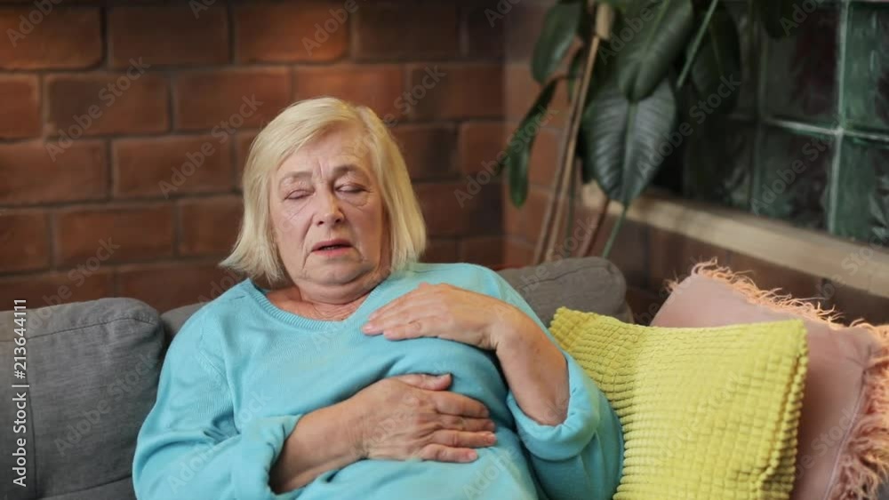 Slavic old woman in blue sweater touching place of heart. Portrait of suffering grandmother having heartache. Indoors. Close-up. Heavy breathing.