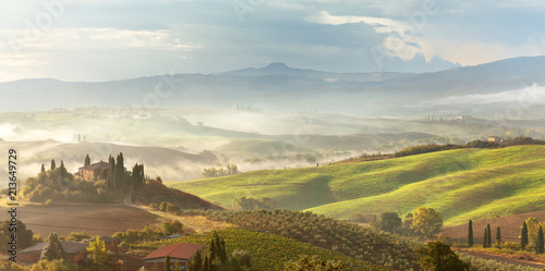 A beautiful Italian traditional rural landscape with autumn fields in the hills and a farmhouse with cypress and olive trees in a foggy morning at dawn photo