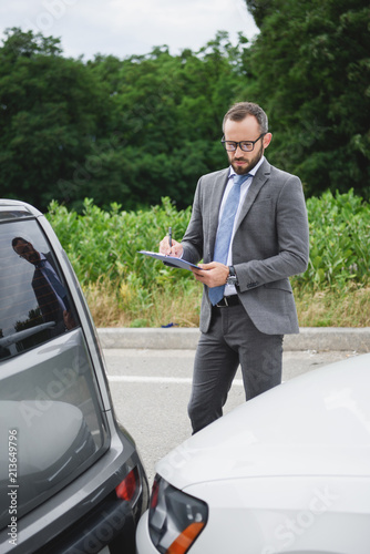 handsome businessman writing something to car insurance after car accident on road