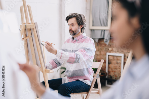 Side view portrait of mature man  painting sitting by easel in art class, enjoying work in spacious sunlit loft studio, copy space