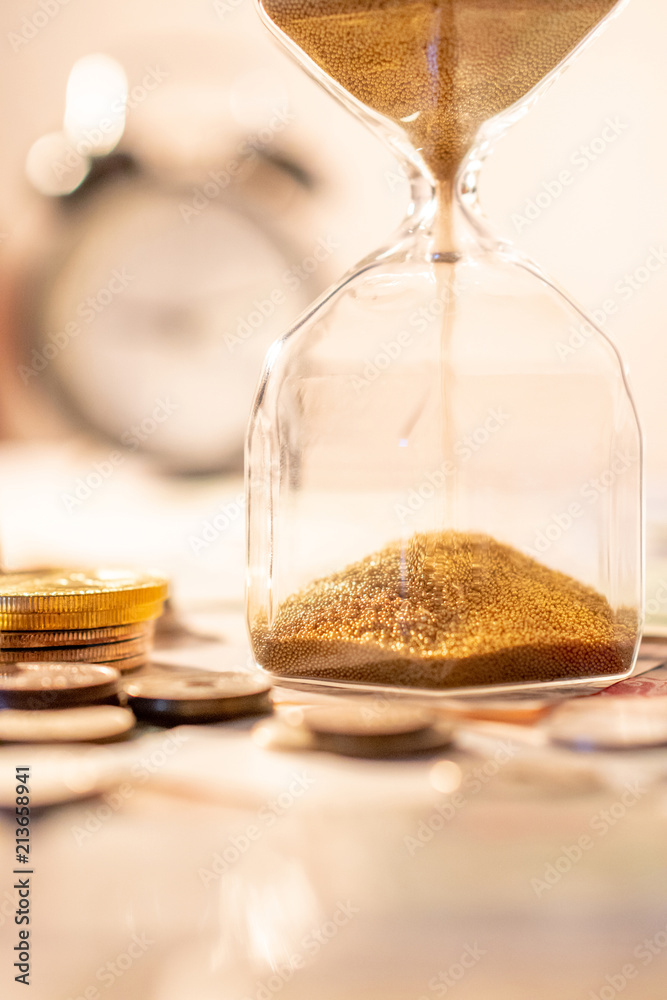 Sand running through the shape of hourglass on table with banknotes and coins of international currency. Time investment and retirement saving. Urgency countdown timer for business deadline concept Sand running through the shape of hourglass on table with banknotes and coins of international currency. Time investment and retirement saving. Urgency countdown timer for business deadline concept