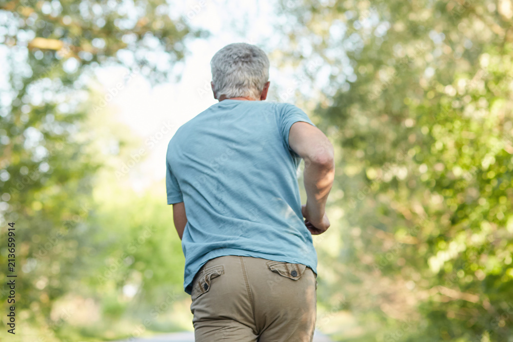 Back view of mature male joggs outside, has morning workout, enjoys sunny day and fresh air, being in motion, has healthy lifestyle. Elderly people, sport, motivation and retirement concept.