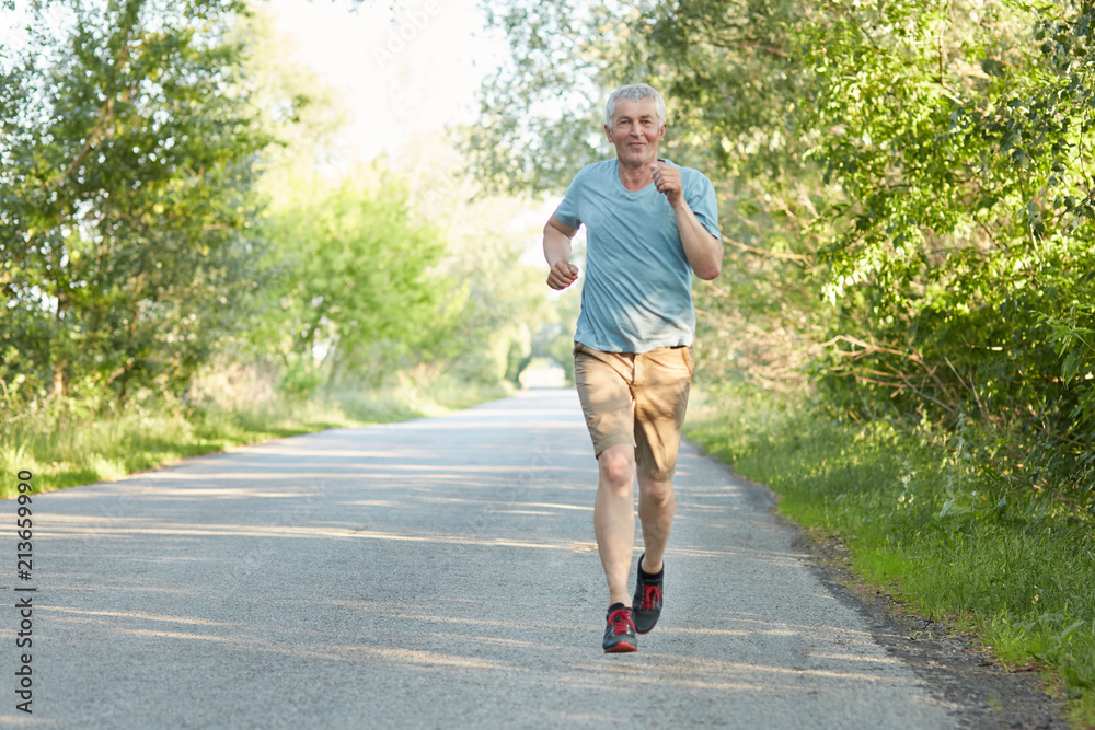 Confident elderly male runner joggs on road, has workout in morning, being  very active, enjoys beautiful nature, being on pension. Pensioner has  outdoor training alone. Sport and motivation concept Stock Photo