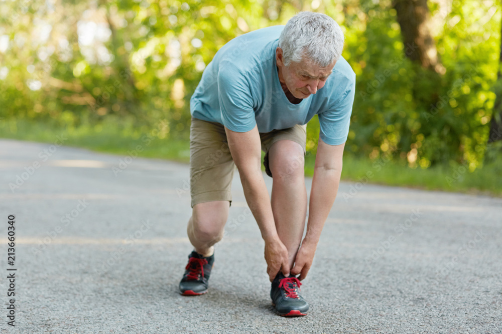 Outdoor view of sporty healthy male pensioner stretches leg, pulled muscle,  dressed in sportswear and sneakers, stands on asphalt, can`t run anymore,  enjoys sunny day and fresh air. Sport concept Stock Photo