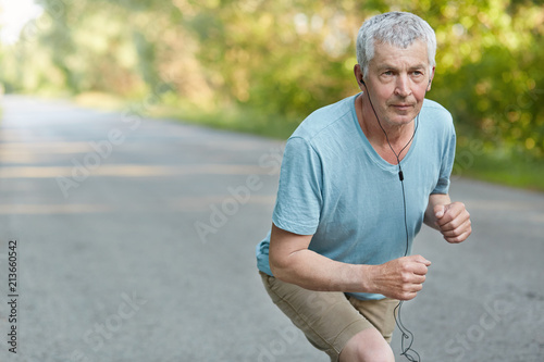 Retired confident male stands on start, ready to take part in sport comeptitions, listens music with earphones and unrecognizable device, has aim to win poses on road in countryside. Healthy lifestyle