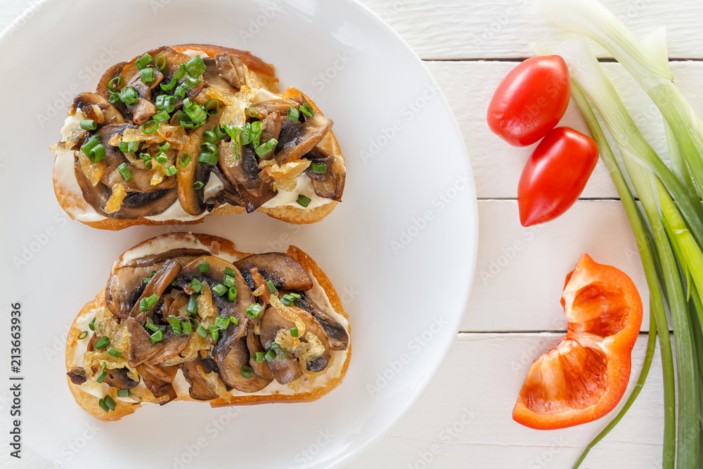 Two fresh bruschettes with mushrooms on lie on a plate next to tomatoes, peppers and onions on a white wooden background. Top view