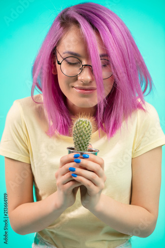 Emotional dyed haired woman with violet hair holding potted plan in hands. Smiling girl wearing spectacles, looking at cactus with happy cheerful expression photo