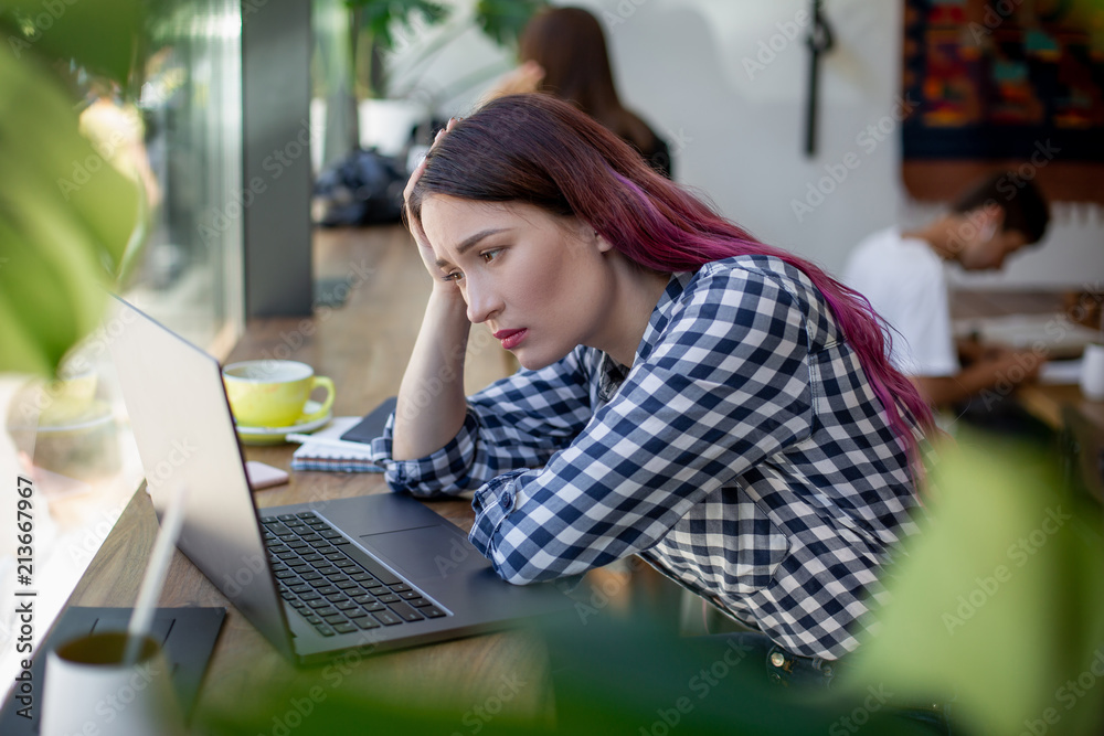 Fototapeta premium Side view of young businesswoman sitting at table in coffee shop. On table cup of coffee and laptop. In background white wall and window.
