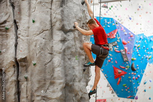 Photo from side of sports guy in red T-shirt training on climbing wall indoors