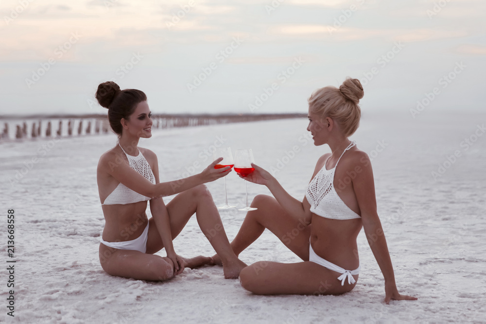 Cheers! Happy two girl friends in sexy bikini having fun on white salty  beach at sunset.