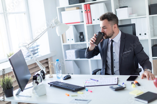 A man sits in the office at the table, drinks coffee and looks at the monitor.