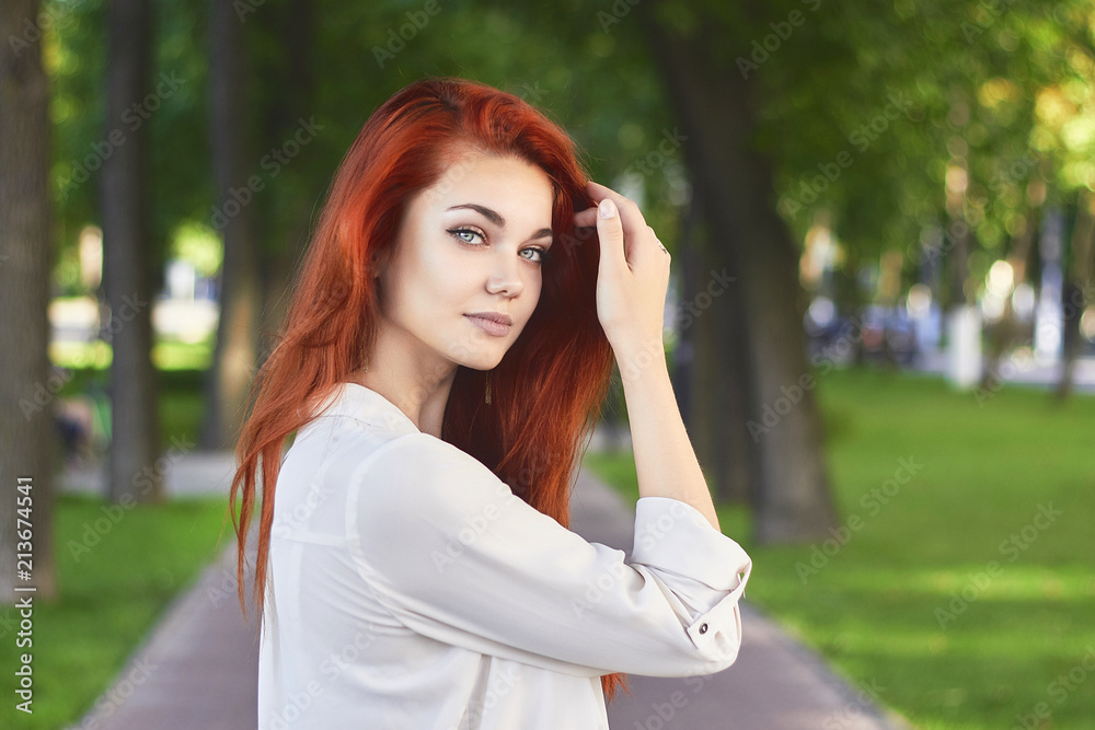 Fototapeta premium Close up of attractive red-haired girl in white shirt with long foxy hair posing in park background. Ginger teenager holds her hand near the head looking at camera.