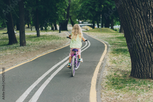 back view of blond child riding bicycle on road in park