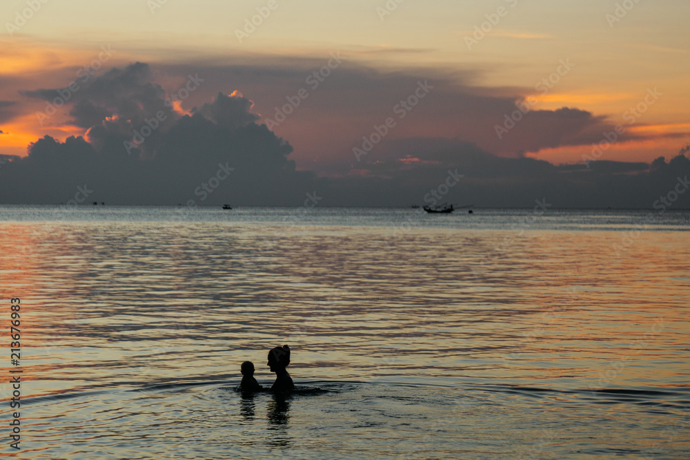 Mother and little child silhouettes in the water on the beach at sunset tropical coast of asia.