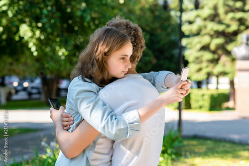 The girl hugs a guy in summer in nature. In the hands of holding smartphones communicates in social networks. Students after school rest. Happy beginning of the relationship. Best friends forever.