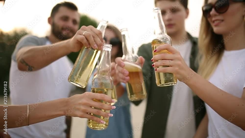 Group of friends having fun enjoying a beverage and relaxing on the beach at sunset in slow motion. Young men and women drink beer standing on a sand in the warm summer evening. Group of friends having fun enjoying a beverage and relaxing on the beach at sunset in slow motion. Young men and women drink beer standing on a sand in the warm summer evening.
