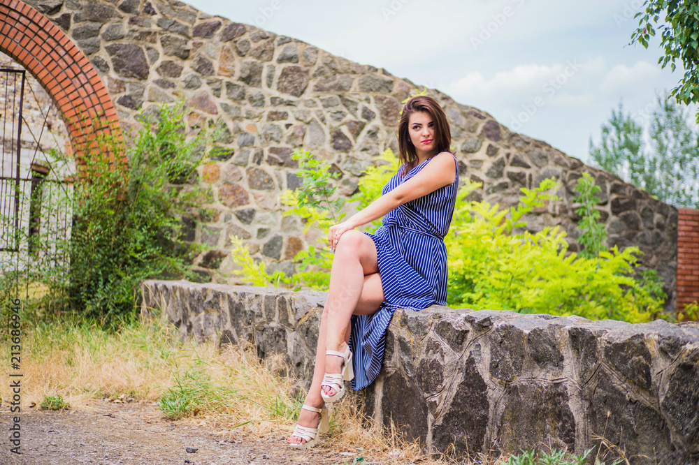 Fototapeta premium The brunette girl in a long blue dress sits on a bard on the background of an arch of red brick and an iron engraved fence with patterns in an abandoned place