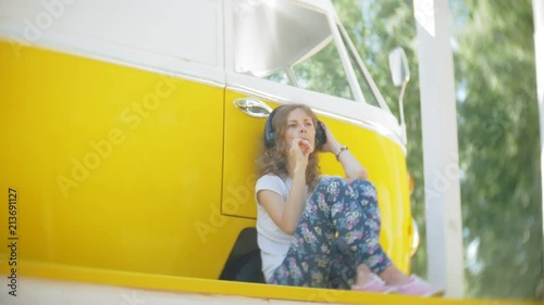 young woman outdoors by yellow vagon car listening music in headphones using smartphone - relaxing, enjoying, concept of technology and travel photo