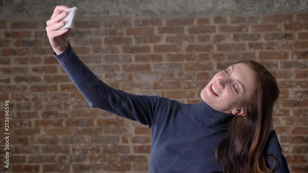 Brown hair girl is making selfie on smartphone, fooling around, communication concept, brick background Brown hair girl is making selfie on smartphone, fooling around, communication concept, brick background