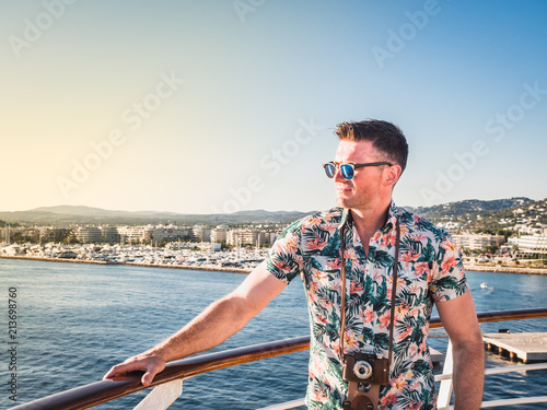 Attractive guy with a vintage camera on the deck of a cruise liner against a background of blue, sea waves, sunset and shoreline. Concept of sea cruises and rest